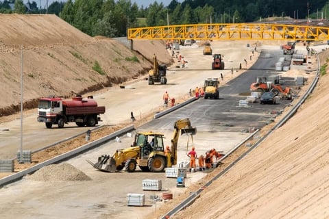 Highway construction site with heavy machinery, workers, and equipment on concrete roadbed under yellow crane and earthen embankments.